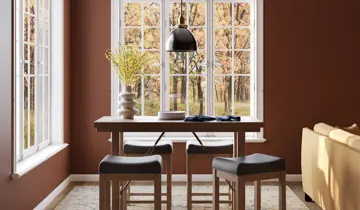 Warm brown dining area with a wooden counter-height table, four black stools, and large windows showcasing autumn trees