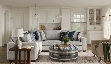 Bright living room with a light gray sectional, blue pillows, round striped ottoman, and white kitchen beyond