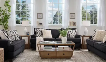 Sunny living room with dark charcoal sofa, armchair, and loveseat, accented by patterned pillows and light wood tables