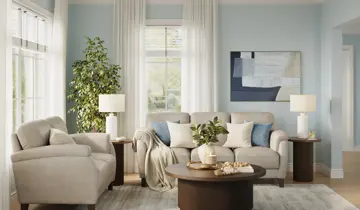 Light blue living room with a beige sofa, armchair, dark wood tables, a green plant, and modern artwork