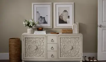 Carved cream console table in a neutral entryway with framed art, flowers, books, and woven basket