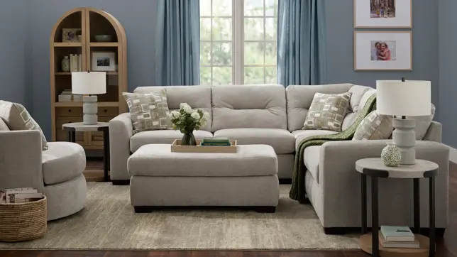 Bright living room with light gray sectional, ottoman, patterned pillows, blue walls, and a wooden bookshelf