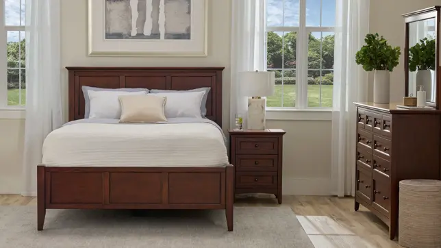 Classic bedroom with dark wood bed, nightstand, and dresser, white bedding, and large windows revealing a green yard
