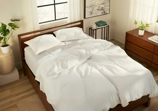 A bedroom with a wooden bed, white bedding, plants, window, and wood furniture