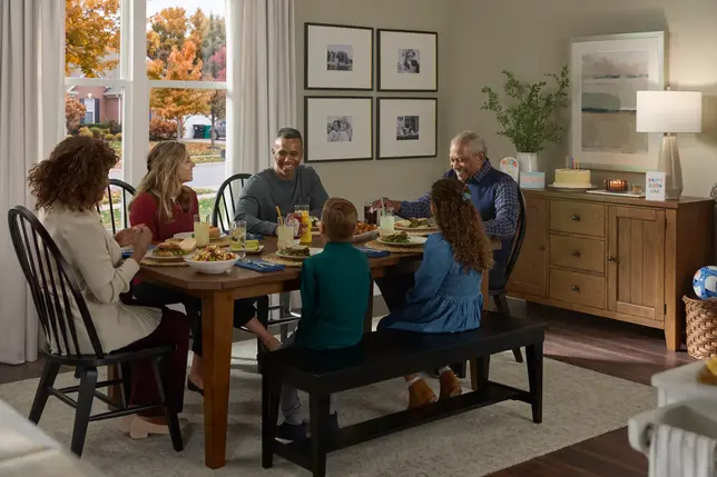 Family dines at a wooden table, surrounded by black chairs and bench, with a wood sideboard and autumn light