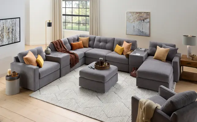Bright, modern living room with a U-shaped gray modular sectional, ottoman, armchair, and orange/yellow pillows on a textured rug