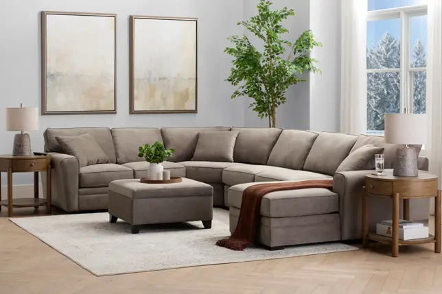 Neutral living room featuring a U-shaped sectional, ottoman, wooden end tables, abstract wall art, and a large plant