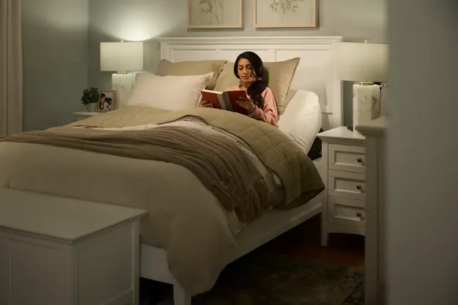 Warm bedroom with white adjustable bed, beige bedding, two nightstands, storage bench, and a woman reading