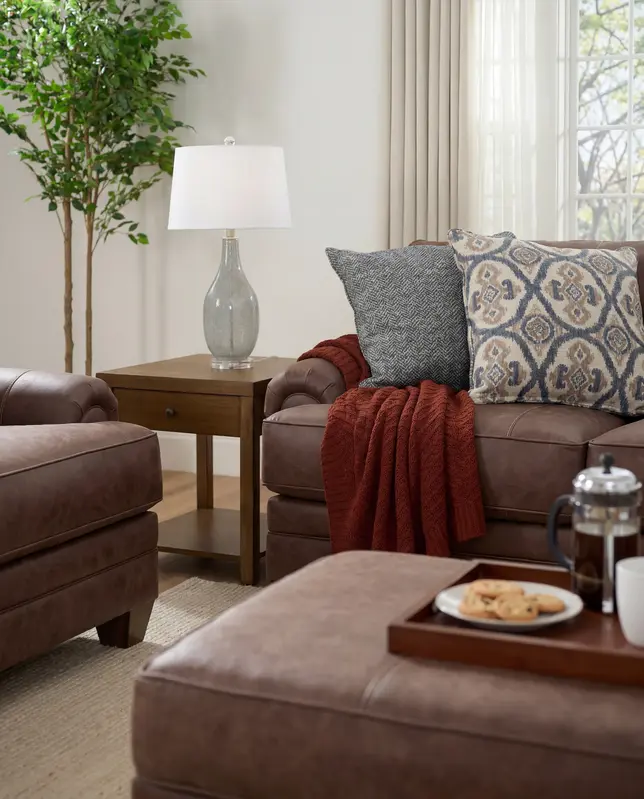 Cozy living room with rich brown leather sofa, armchair, and ottoman, featuring a wooden end table and table lamp