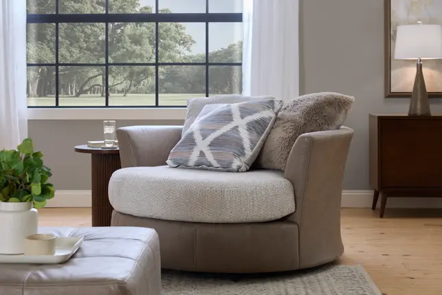 Inviting living room with a round taupe swivel chair, patterned pillow, wooden side table, and large window
