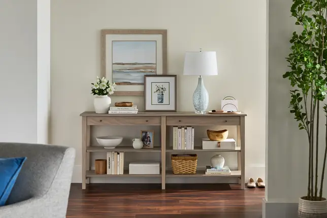 A neutral living room features a long, light gray console table with open shelving, styled with books, decor, and art