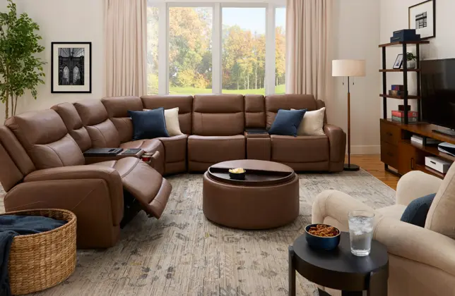 Spacious living room with brown leather reclining sectional, built-in consoles, round ottoman, and autumn window
