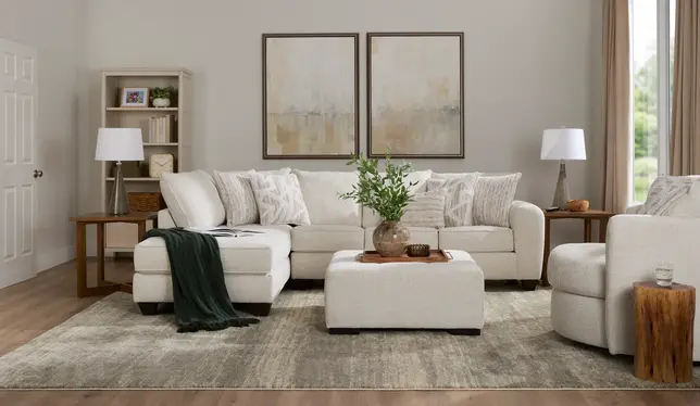 Neutral living room with cream sectional, ottoman, armchair, abstract art, and a bookshelf on a patterned rug