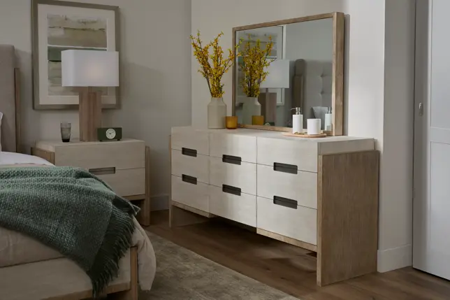 Serene bedroom with light wood dresser, mirror, nightstand, bed, and soft green throw blanket