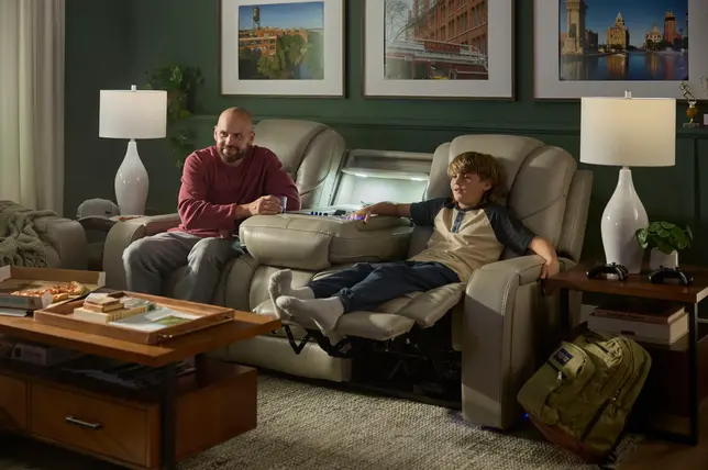 A father and son relax on a gray reclining sofa with a center console, coffee table, and green walls