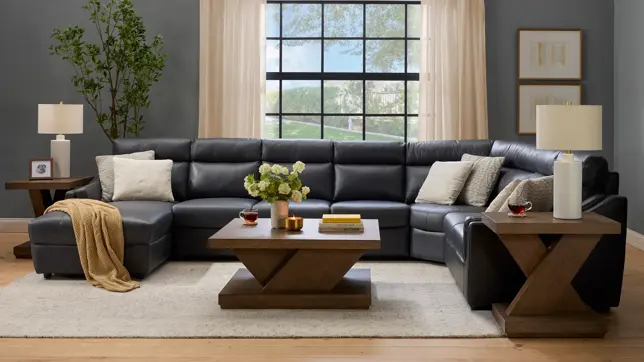 A modern living room features a dark gray sectional sofa, geometric wooden tables, and natural light from a large window