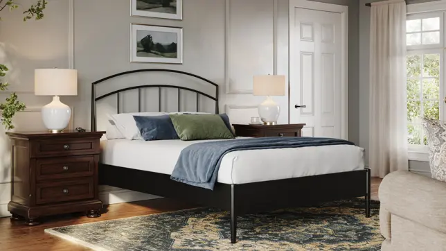 A bedroom with a black metal bed, dark wood nightstands, white lamps, a patterned rug, and natural light