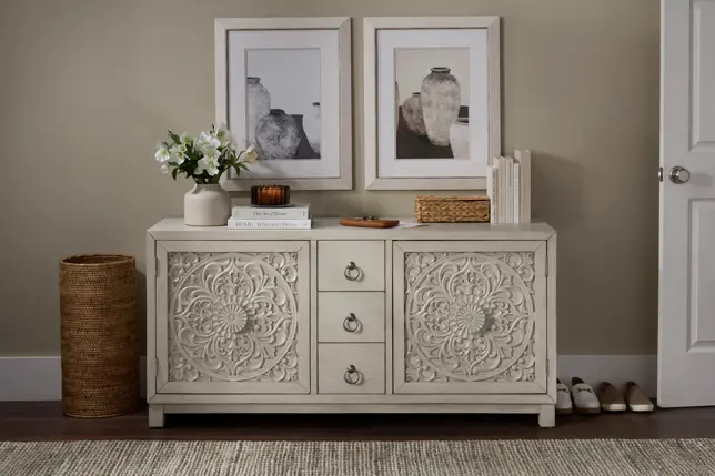 Entryway styled with a light carved sideboard, two framed prints, flowers, and a natural woven basket