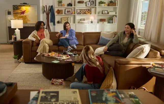 A warm living room features a large brown leather sectional, round coffee table, and friends relaxing
