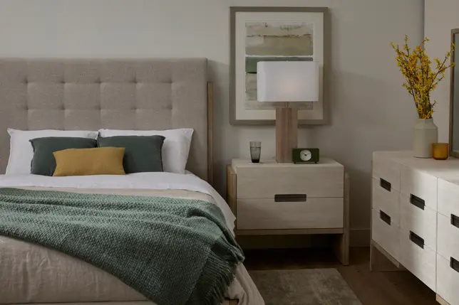 Neutral bedroom with upholstered bed, nightstand, dresser, lamp, and framed art